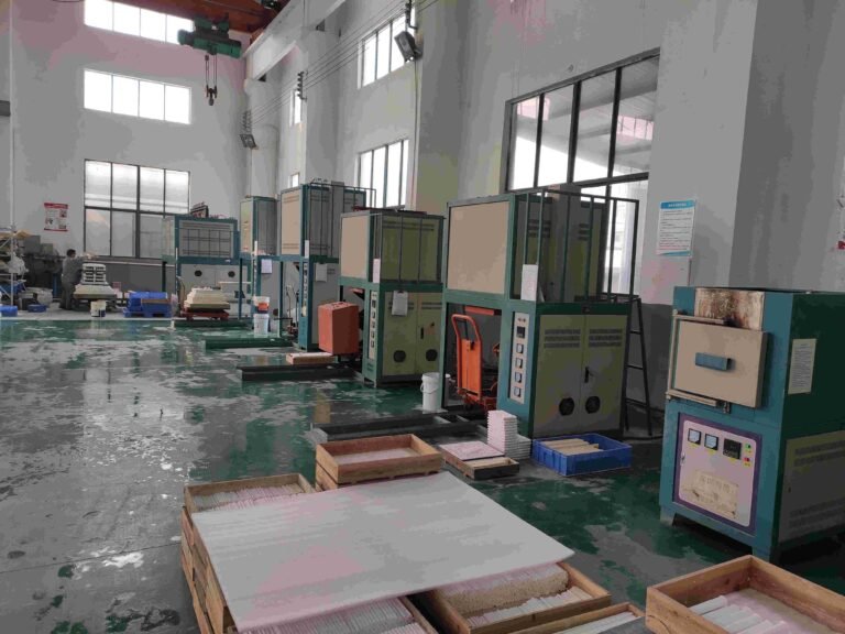 A row of firing furnaces down the length of the bay, loaded setter boards stacked on pallets in the foreground